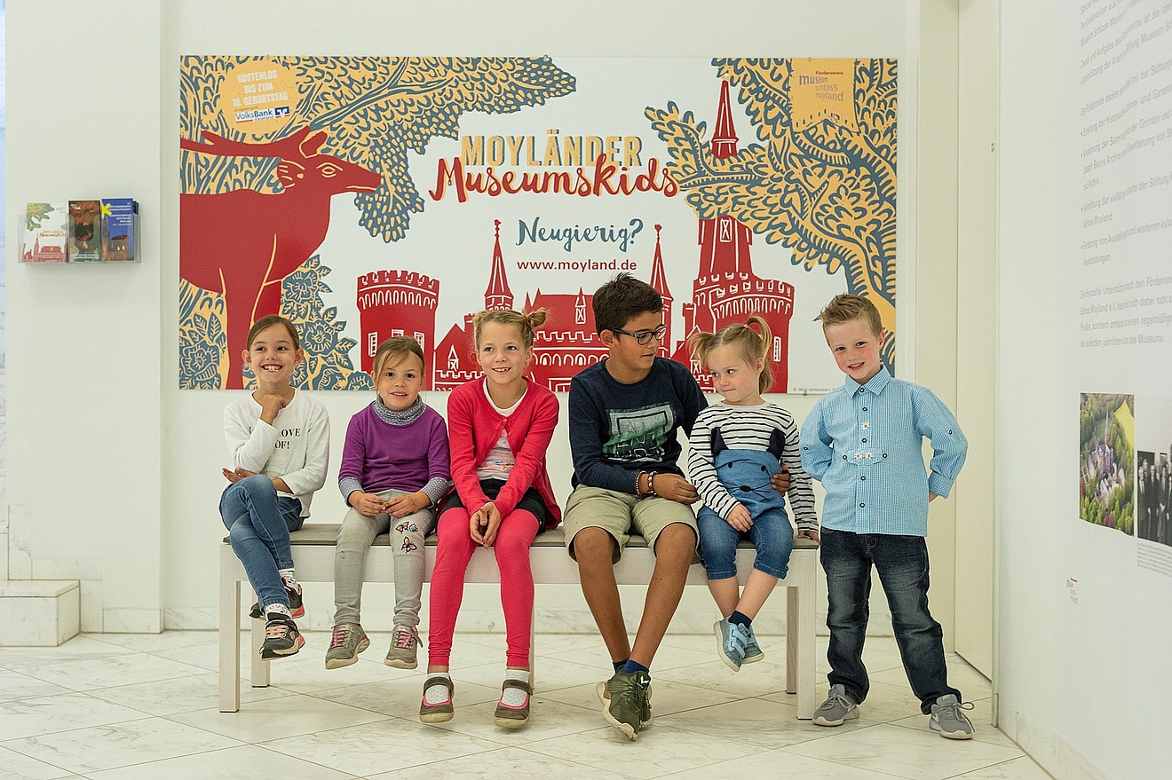 Kinder sitzen auf Bank, im Hintergrund Plakat Moyländer Museumskids